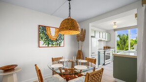 A modern kitchen with white cabinets, a dining area with a glass table and wicker chairs, and a view of greenery outside. - Kauai The Cliffs At Princeville 4209 1 Bedroom Condo (Princeville)