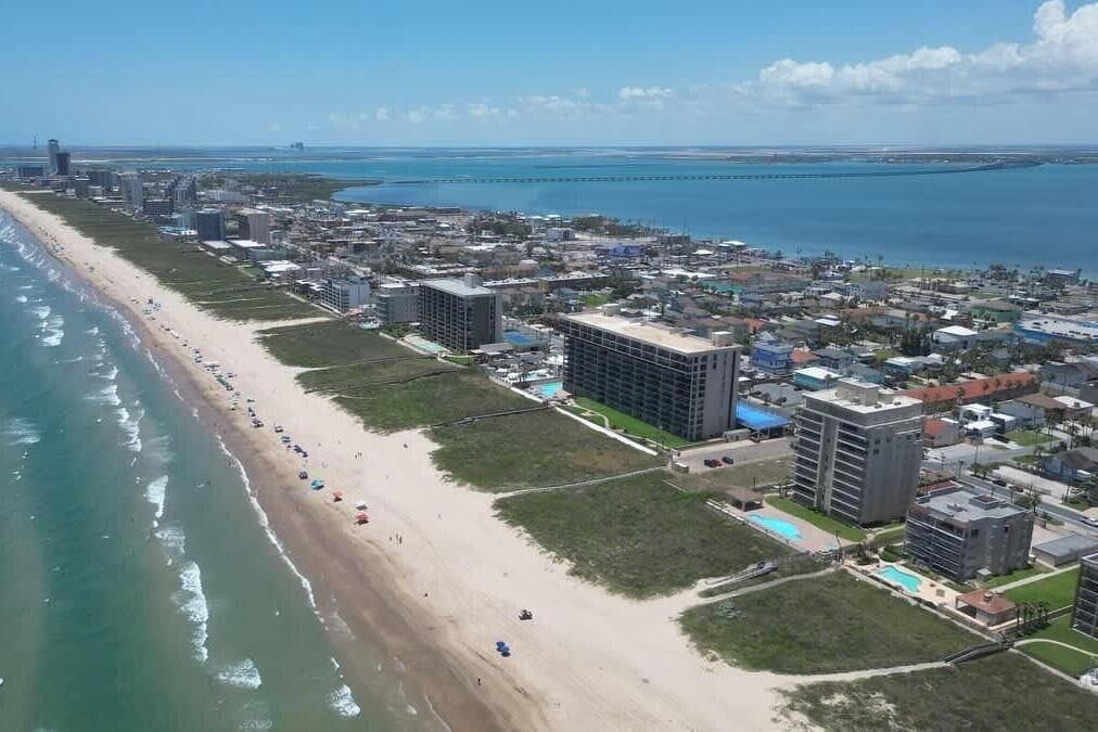 An aerial view of a coastal city with a long beach, buildings, and a bridge in the distance.