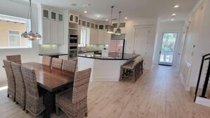 A modern kitchen with white cabinets, a central island, and a dining area with wicker chairs. - Padre Beach House 5 Bedroom Home by RedAwning (South Padre Island)