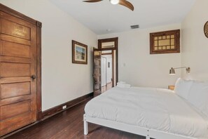 Een slaapkamer met een houten deur, een bed met witte beddengoed, een plafondventilator en een raam met houten kader.