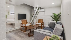 A compact living space with a sofa, two wooden chairs, a small table, a TV, and a staircase leading to an upper level.