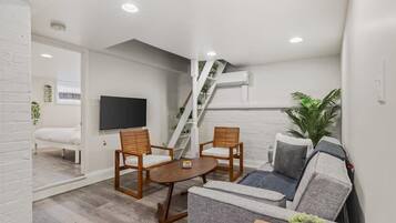 A modern living room with a sofa, two wooden chairs, a coffee table, a flat-screen TV, and a staircase with plants.