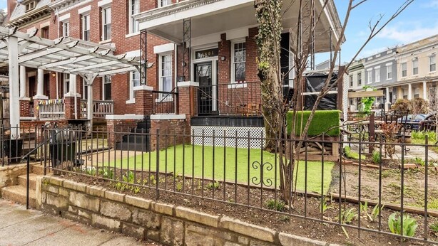 A two-story brick house with a front porch, a small front yard with a lawn, and a metal fence.