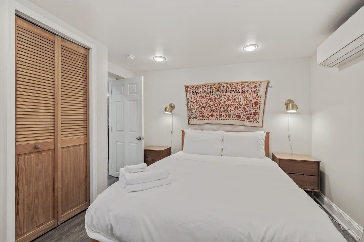 A bedroom with a large bed, wooden wardrobe, two bedside tables with lamps, and a patterned tapestry on the wall.