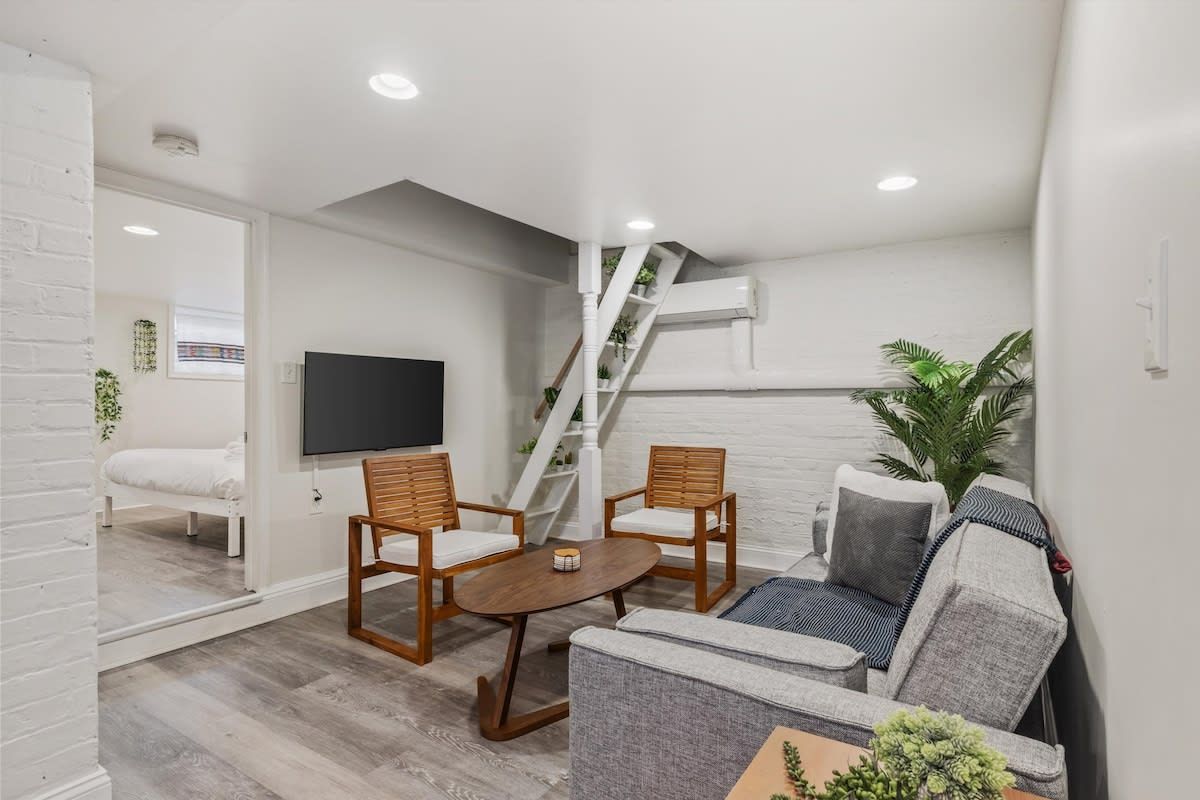A modern living room with a sofa, two wooden chairs, a coffee table, a flat-screen TV, and a staircase with plants.