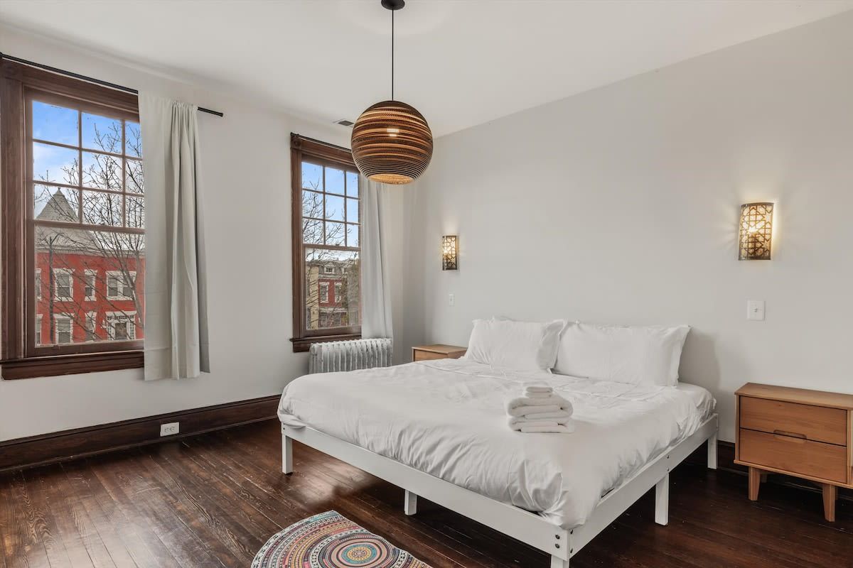A bedroom with a white bed, wooden floor, a hanging pendant light, a window with curtains, and a view of buildings outside.