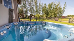 Un jacuzzi en un patio trasero con terraza de madera, una valla y árboles al fondo.
