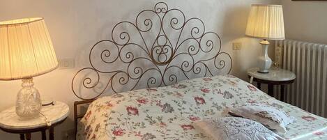 A neatly made bed with floral bedding, two bedside lamps, a framed cityscape picture, and a radiator.