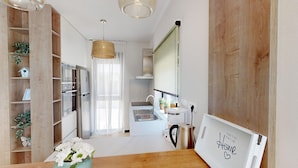 A modern kitchen with a built-in oven, refrigerator, and sink. There is a wooden shelf unit with plants, a hanging light fixture, and a window with sheer curtains. - Villa Silvia in Mihatovici (Mihatovici)