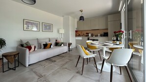 A modern living room with a sofa, chairs, and a dining table. There is a kitchen in the background with cabinets and appliances.