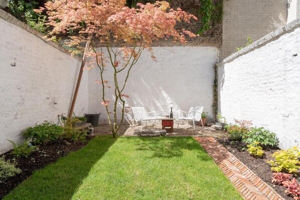 A small backyard with a lawn, a brick pathway, and a seating area with white chairs.