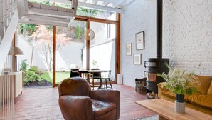 A modern living room with a leather chair, a wooden coffee table, and a sofa. There is a fireplace and a dining area visible through large windows. - Architect Garden House (Brussels)