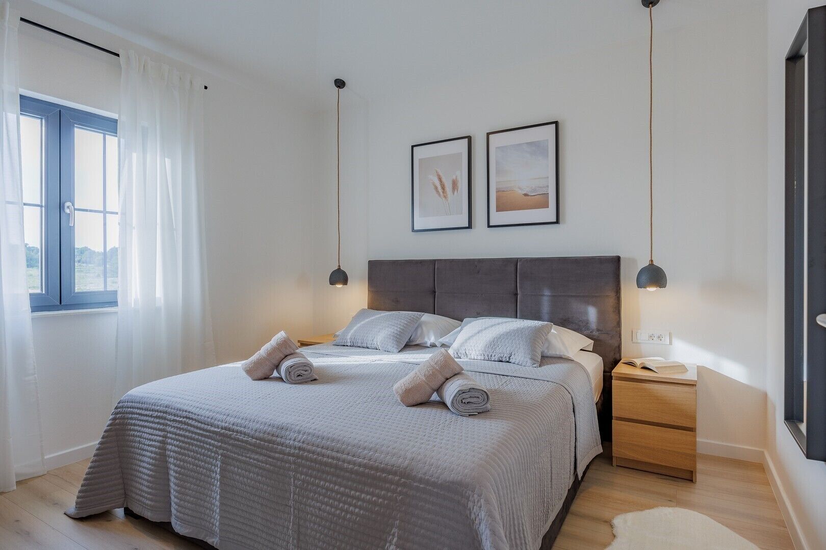 A neatly made bed with a grey bedspread and pillows, a bedside table with a lamp, and two framed pictures on the wall.