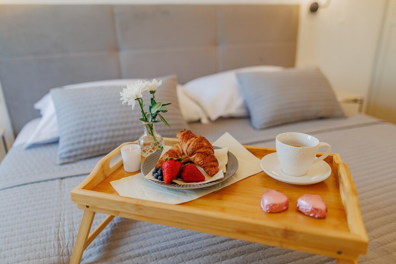 A bed with a wooden tray holding a croissant, strawberries, a cup of coffee, and pink macarons.
