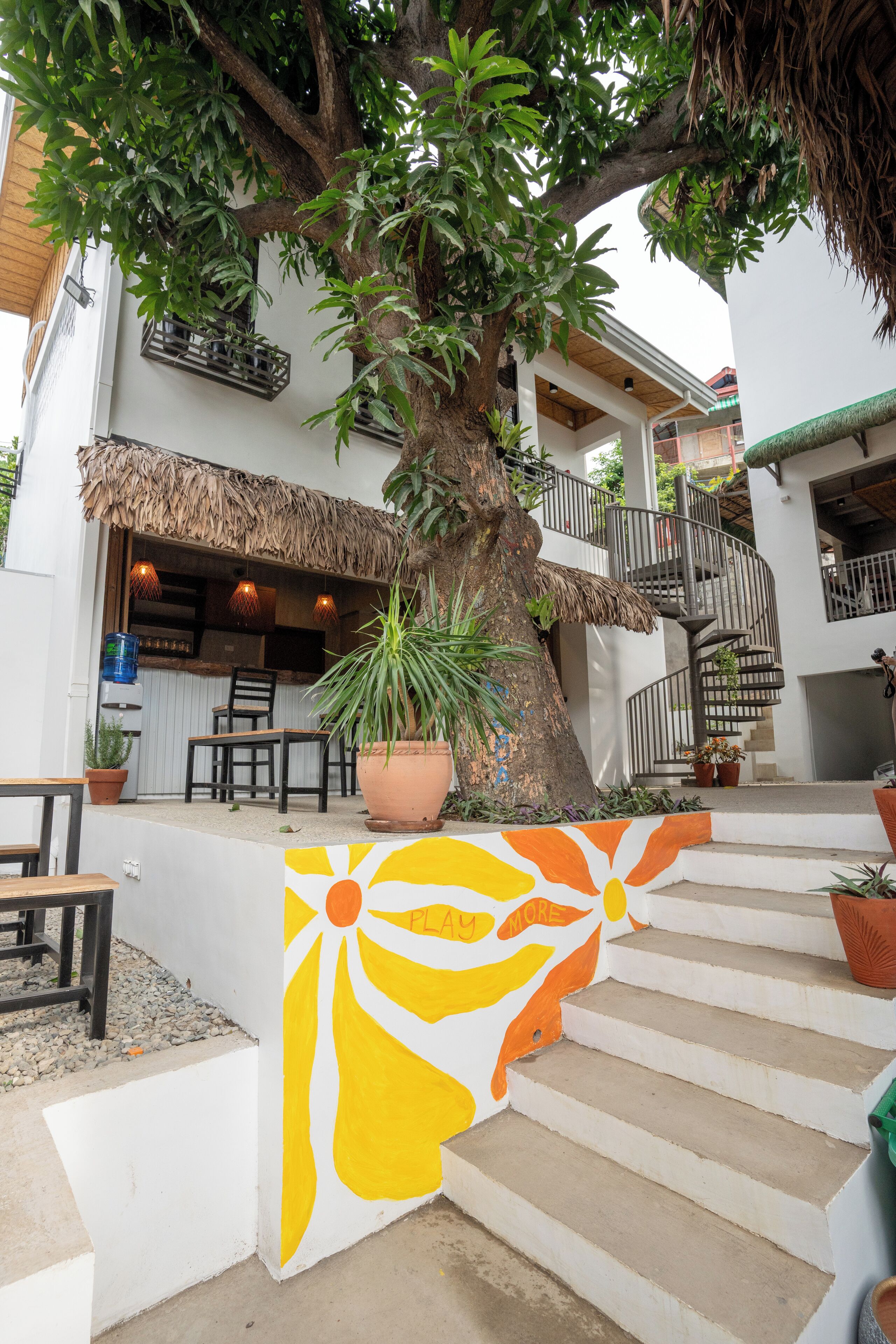 A staircase with a colorful mural, a tree, and a building with a thatched roof and outdoor seating.