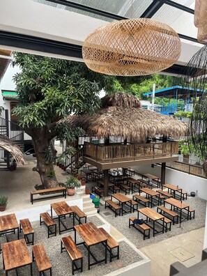 An indoor seating area with wooden tables and benches, a large tree, and a thatched-roof structure. - The Circle Hostel (San Juan)