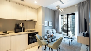 Cocina moderna con estufa a gas, microondas y cafetera. Hay una zona de comedor con una mesa redonda y dos sillas. Se ve un sofá y un sillón colgante cerca de un amplio ventanal con vistas a edificios.