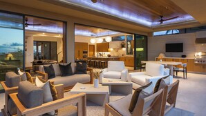 A spacious living area with a large sofa, coffee table, and chairs, a kitchen with a TV, and a view of the outdoors. - Upscale 6BR One Ocean Estate GE3 at Mauna Lani – Golf Course Views & Pool (Kamuela)