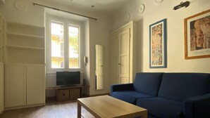 Olohuoneessa on sininen sohva, puinen sohvapöytä, jalustalla oleva televisio ja ovi, joka johtaa toiseen huoneeseen.