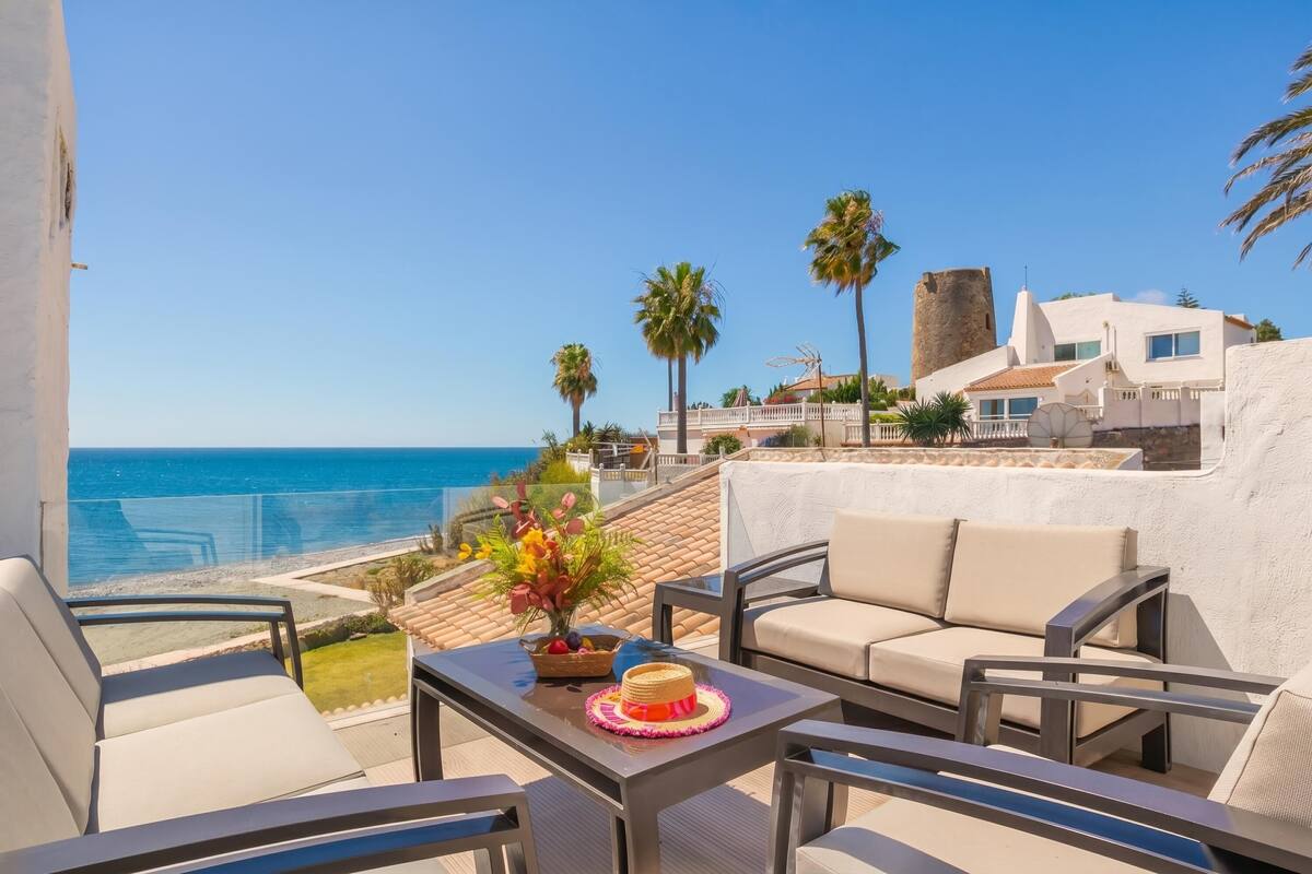 A balcony with a view of the sea, outdoor furniture, and a decorative plant arrangement.