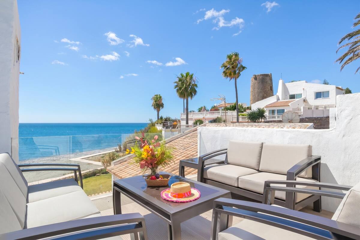 A balcony with outdoor furniture, a view of the sea, and a building with a tower.