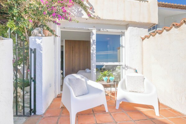A small patio with two white chairs and a table, a sliding glass door leading to a room, and a small garden area with plants and a pink flowering bush. - Bahia Dorada Beach (Estepona)