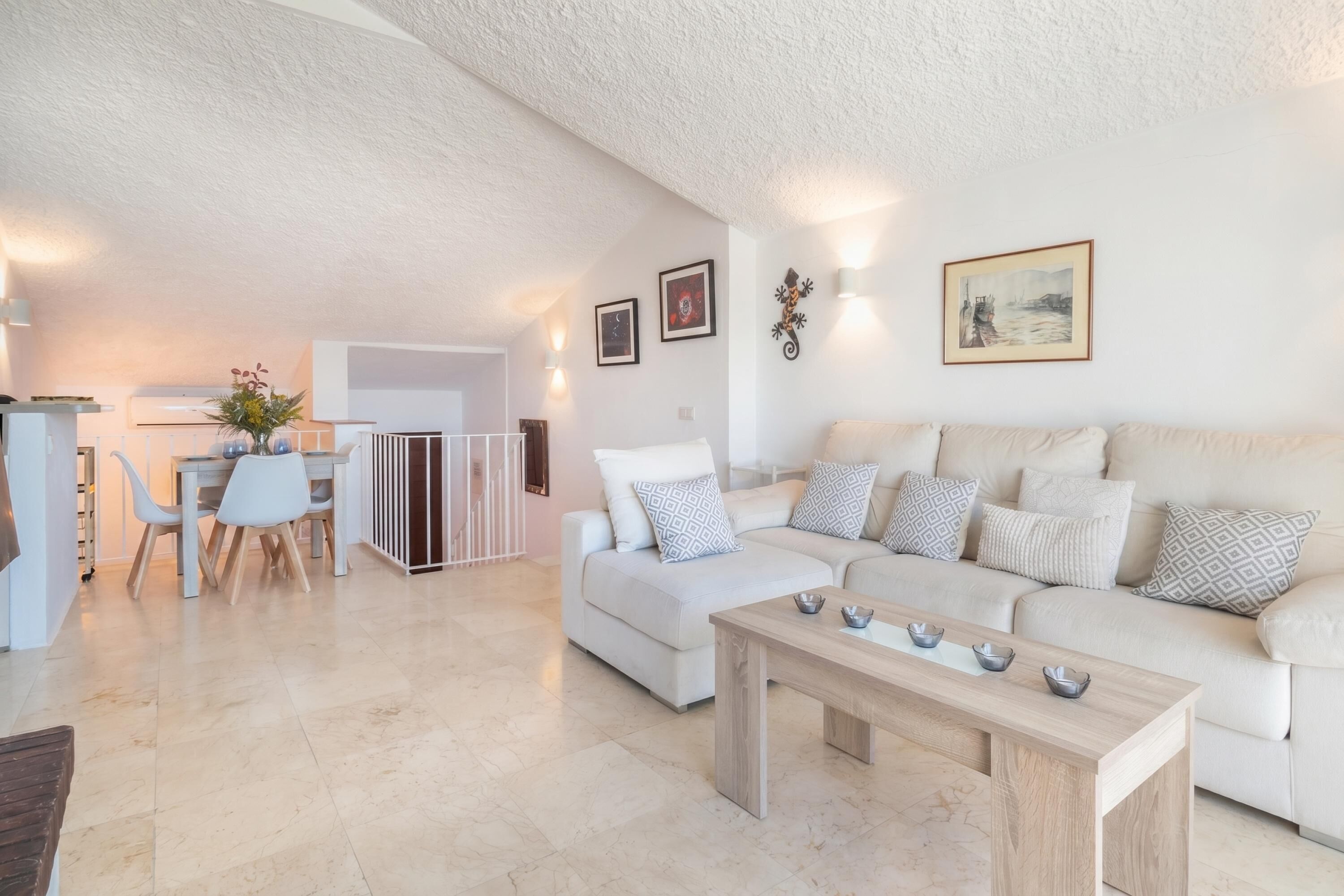 Ein modernes Wohnzimmer mit einem weißen Sofa, einem Holztisch und einem Essbereich mit weißem Tisch und Stühlen.