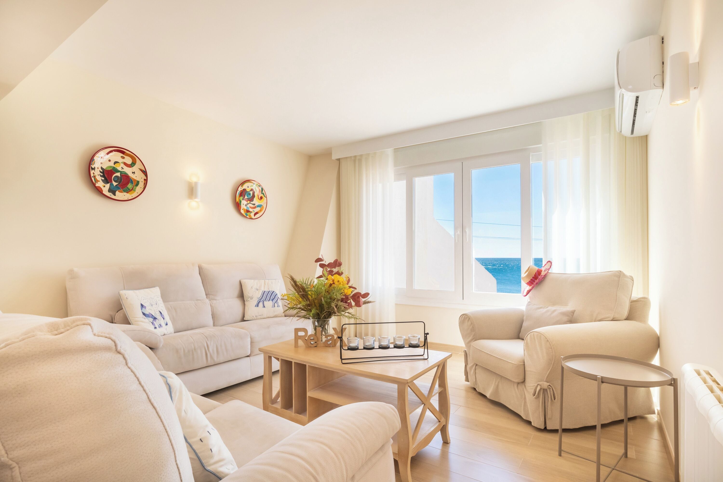 A modern living room with a sofa, armchair, and coffee table, a view of the sea through large windows, and decorative wall art.
