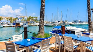 A marina with boats docked, palm trees, and outdoor seating with blue tables and white chairs. - Bahia Dorada Beach (Estepona)