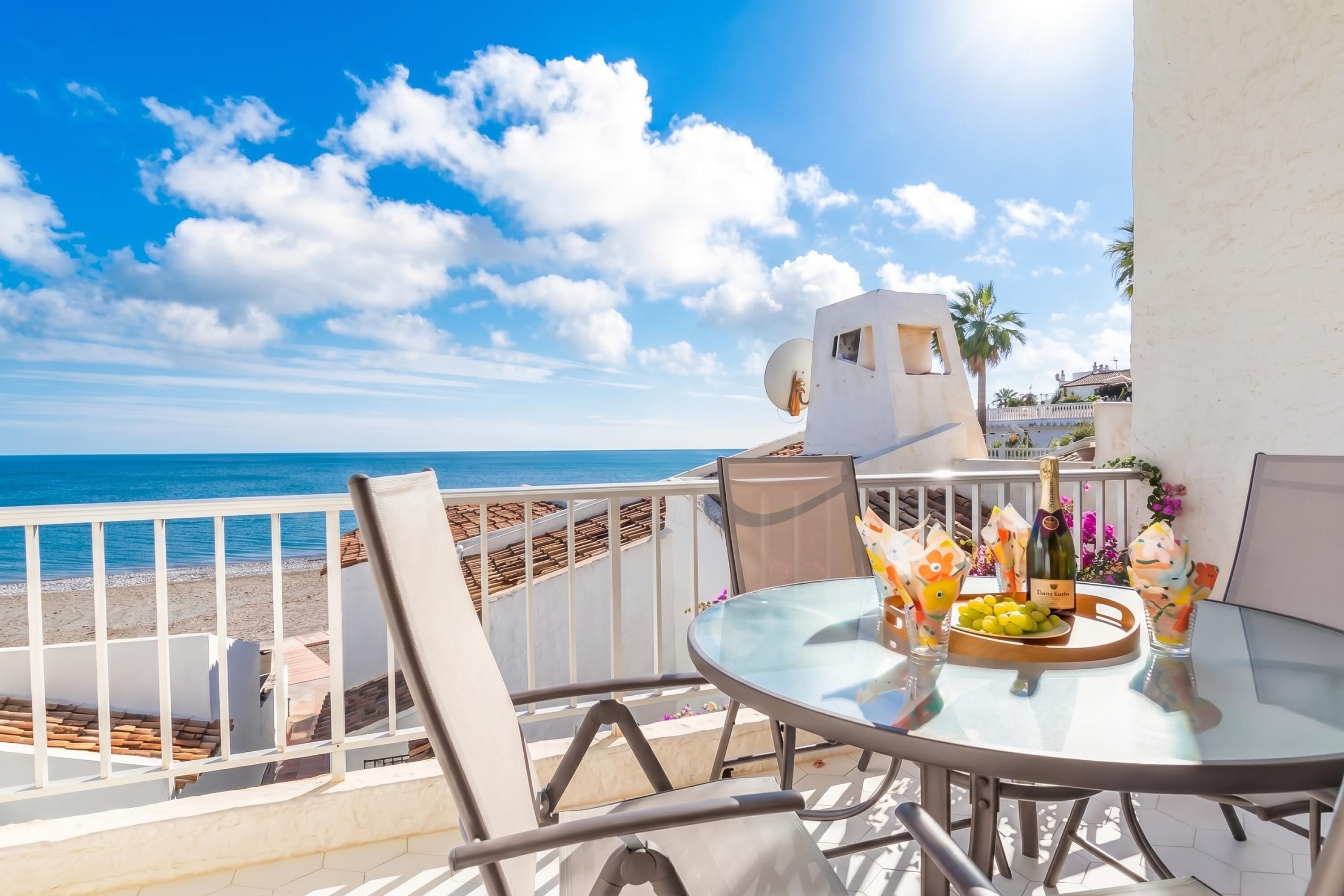 A balcony with a glass table, chairs, and a view of the beach and ocean.