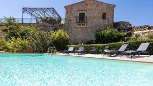 Ein Schwimmbad mit Liegestühlen, ein Steinhaus und eine Metallkonstruktion im Hintergrund.