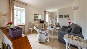 Ein großzügiger Wohnraum mit grauem Sofa, weißen Korbstühlen und einem kleinen Tisch. Es gibt einen Essbereich mit Tisch und Stühlen, und im Hintergrund befindet sich eine Küche.