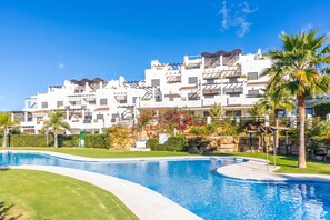 Une piscine à bord incurvé, entourée d’une pelouse bien entretenue et de palmiers. Au fond, on aperçoit un immeuble à plusieurs étages, doté de balcons et d’un mur en pierre.
