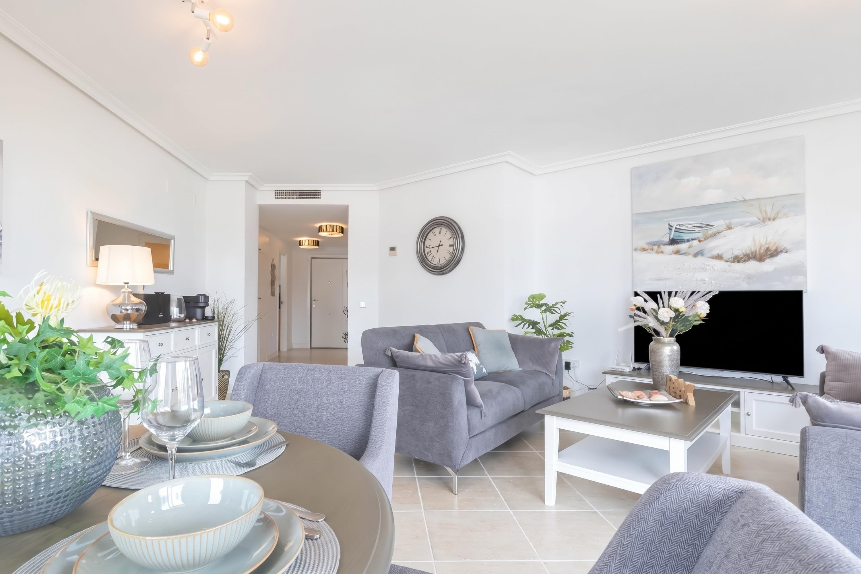 Un salon moderne avec un canapé gris, une table à manger dressée avec des assiettes et des verres, un téléviseur à écran plat et un tableau représentant un paysage enneigé.