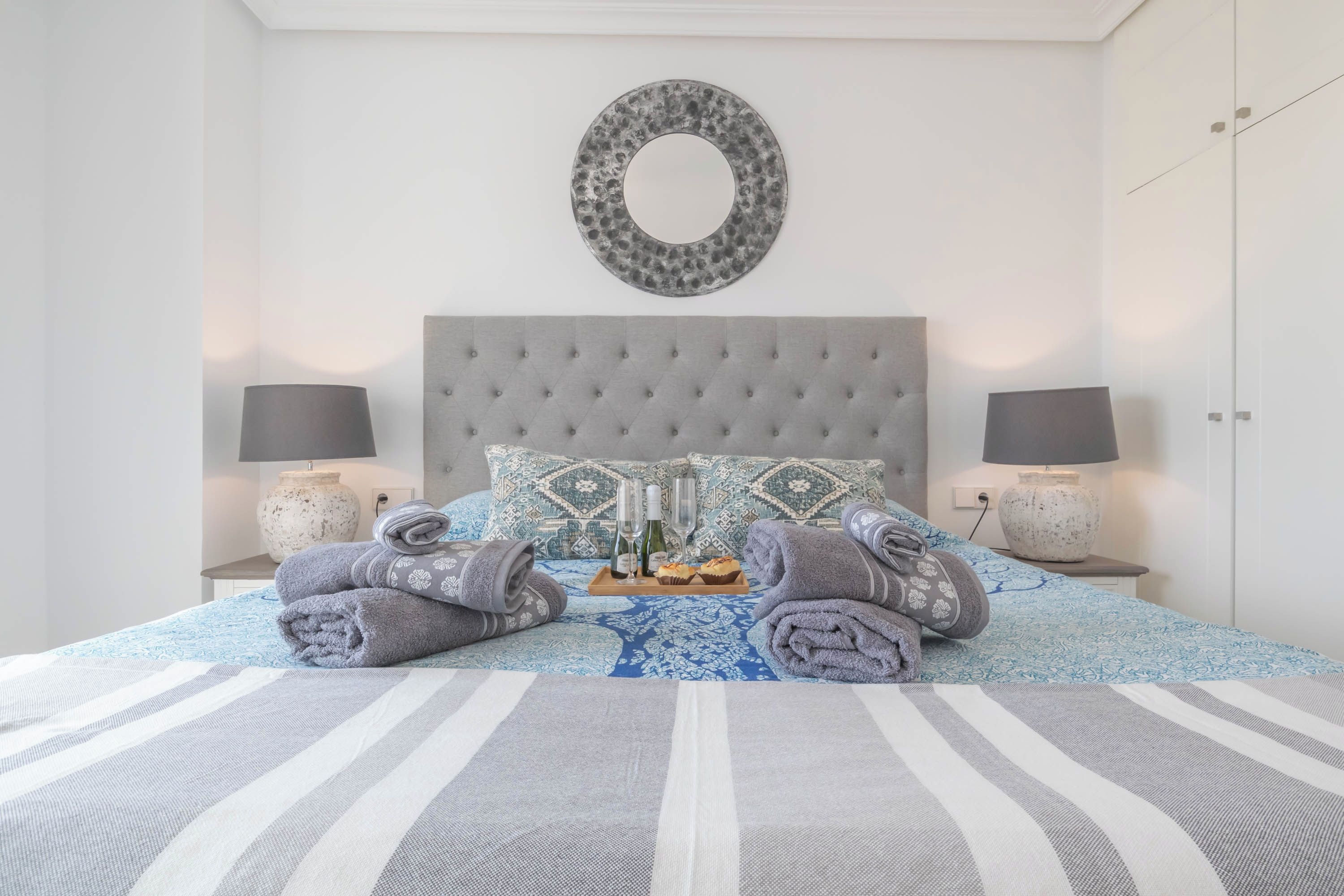 Een opgemaakt bed met een gestreept dekbedovertrek, twee kussens en een ronde spiegel aan de muur.