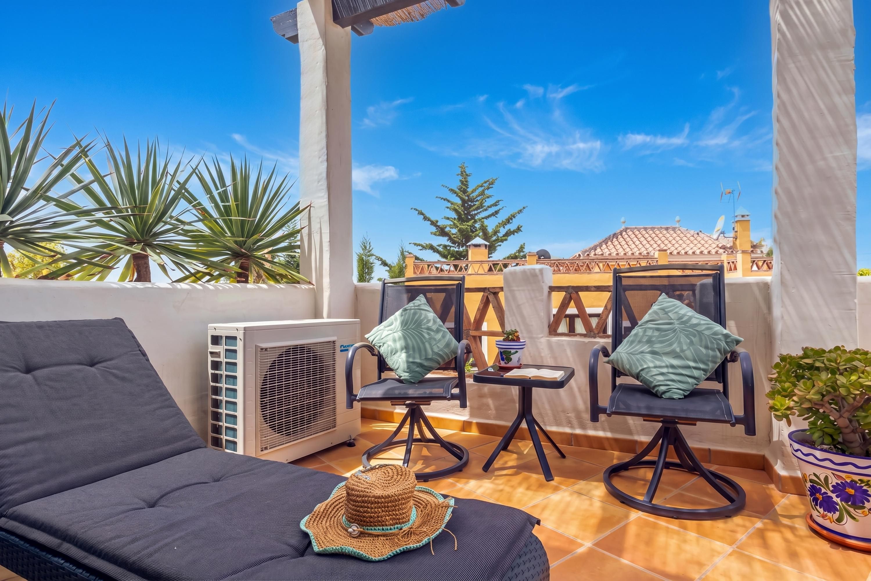 A balcony with a sofa, two chairs, a small table, a potted plant, and an air conditioning unit.
