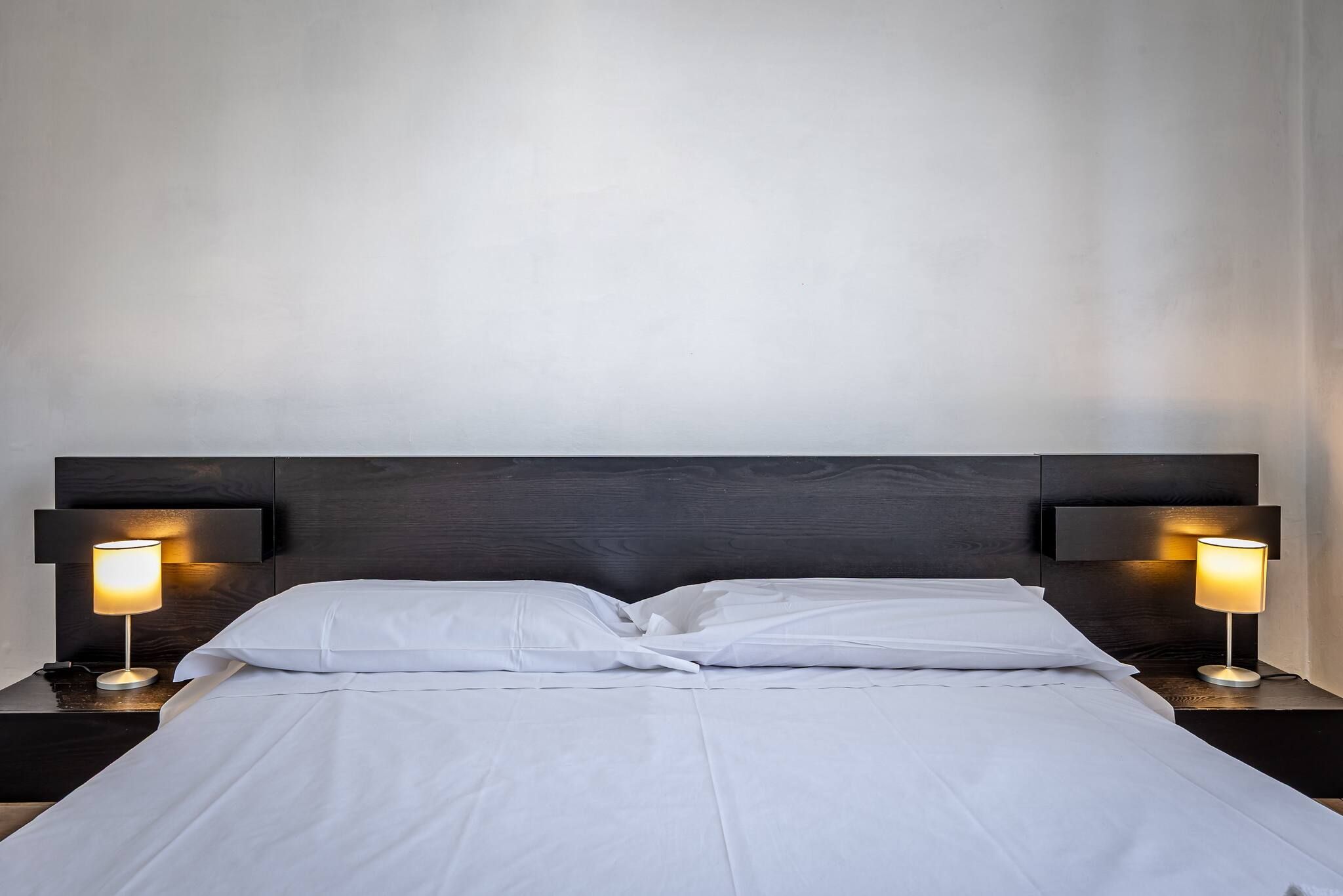 A neatly made bed with white linens, flanked by two bedside tables with lamps, against a plain wall.