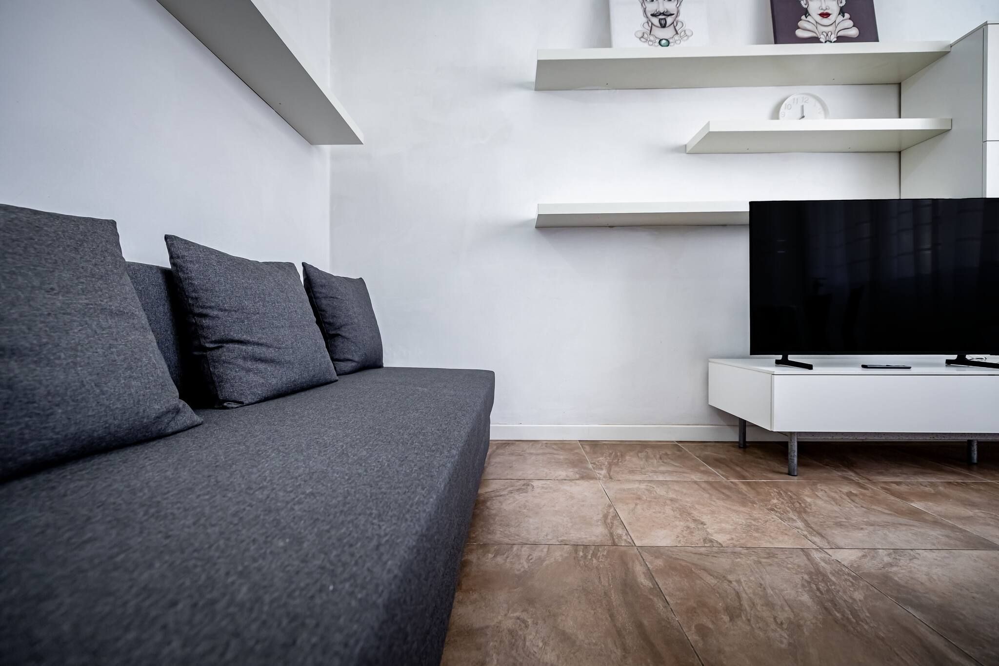A modern living room with a grey sofa, a flat-screen TV on a white stand, and a wall-mounted shelf with decorative items.