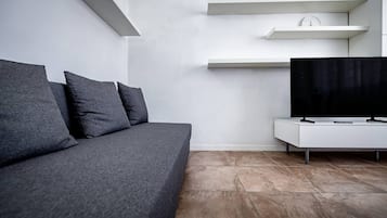 A modern living room with a grey sofa, a flat-screen TV on a white stand, and a wall-mounted shelf with decorative items.