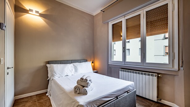 Una cama bien tendida con toallas enrolladas, un cabecero gris y una ventana con persianas.