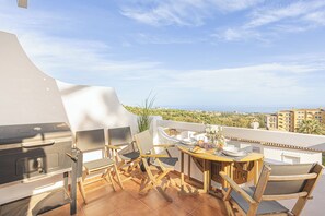 Una terraza con parrilla, mesa de comedor, sillas y vistas al mar y al follaje.