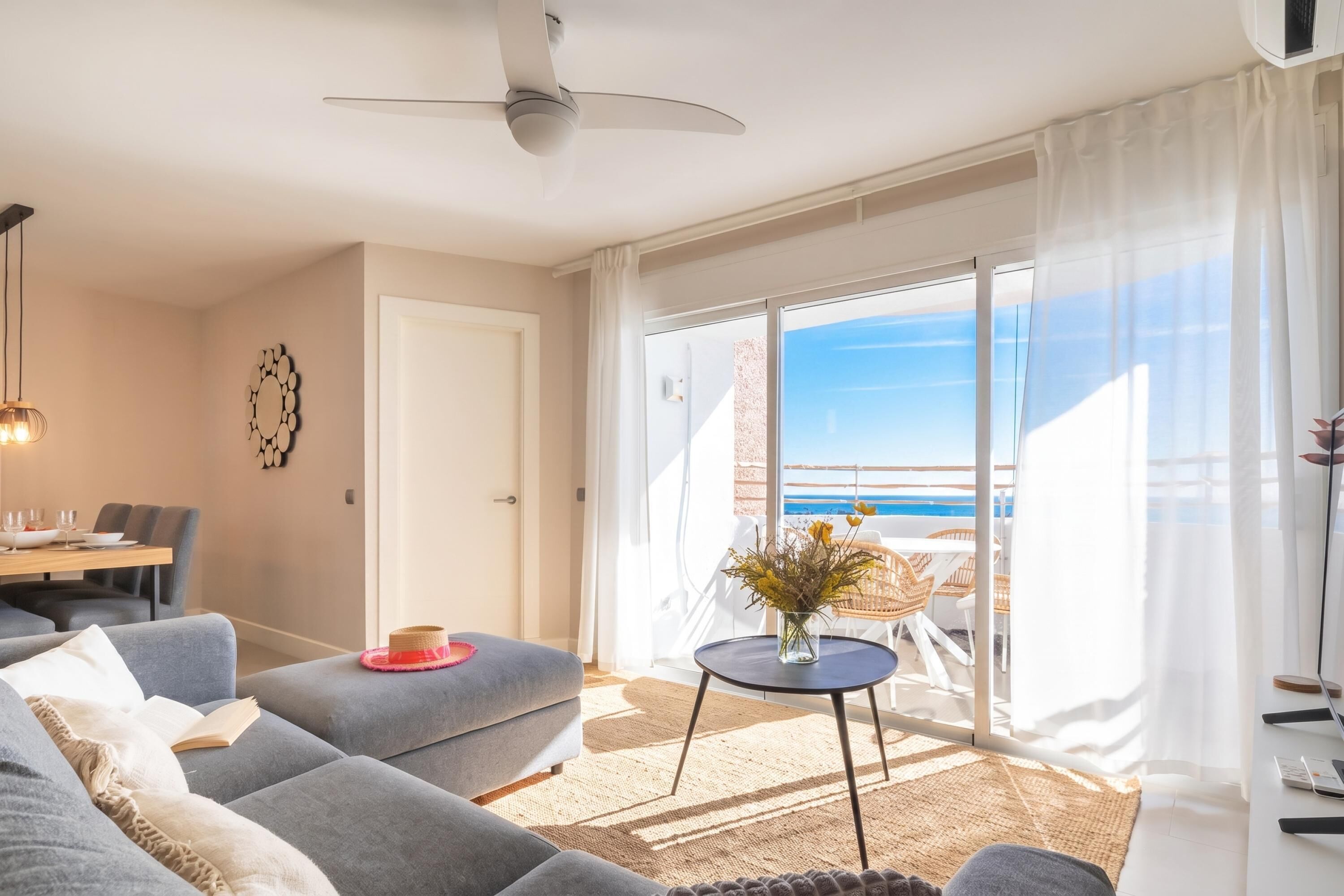 A modern living room with a sofa, a dining table, and a balcony with a view of the sea.