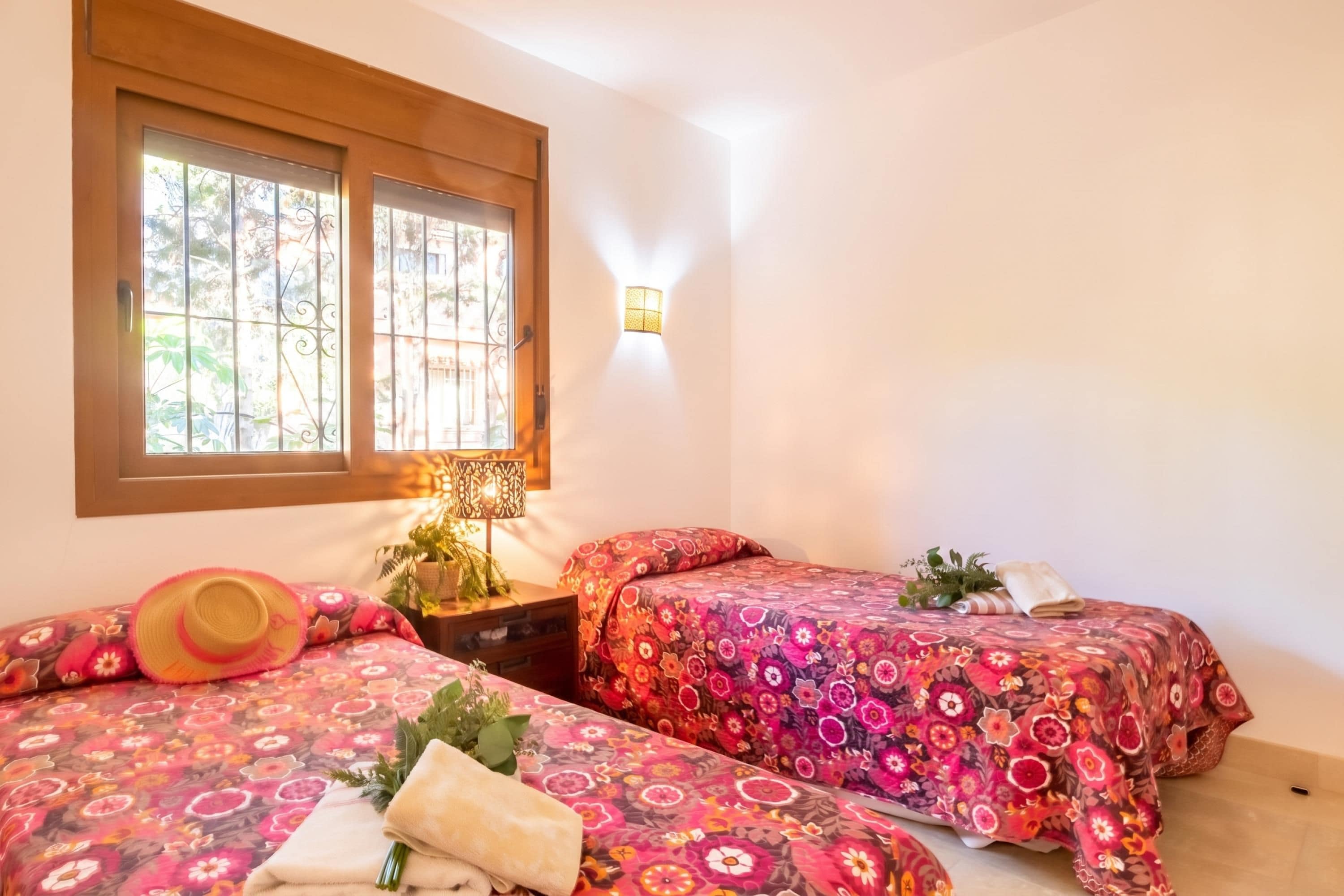 A bedroom with two beds, a window, a wall-mounted lamp, and a nightstand with a lamp.