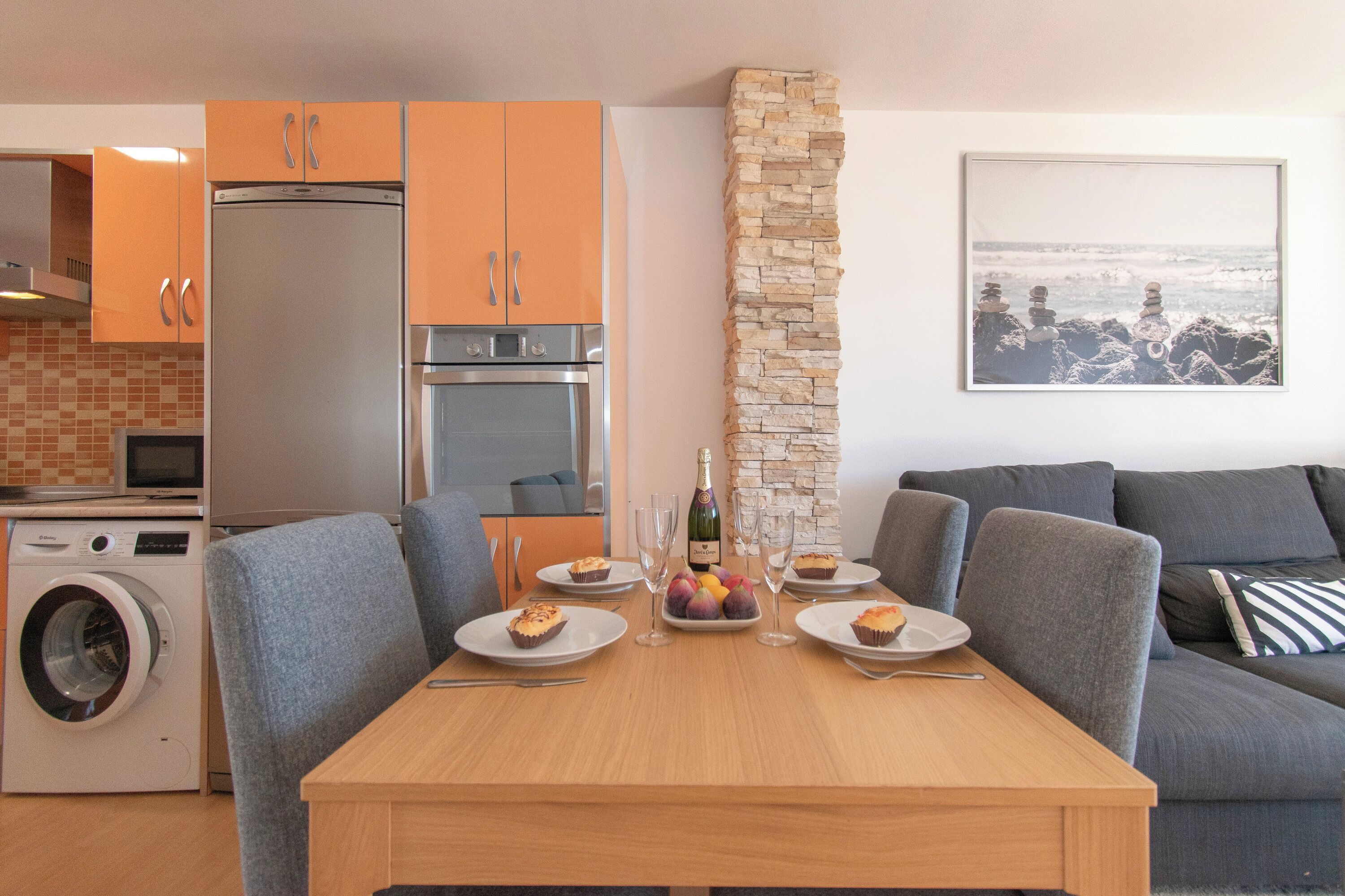 Une cuisine moderne avec des meubles de couleur orange, un mur d’accent en pierre et un coin repas avec des chaises à revêtement gris et une table en bois.