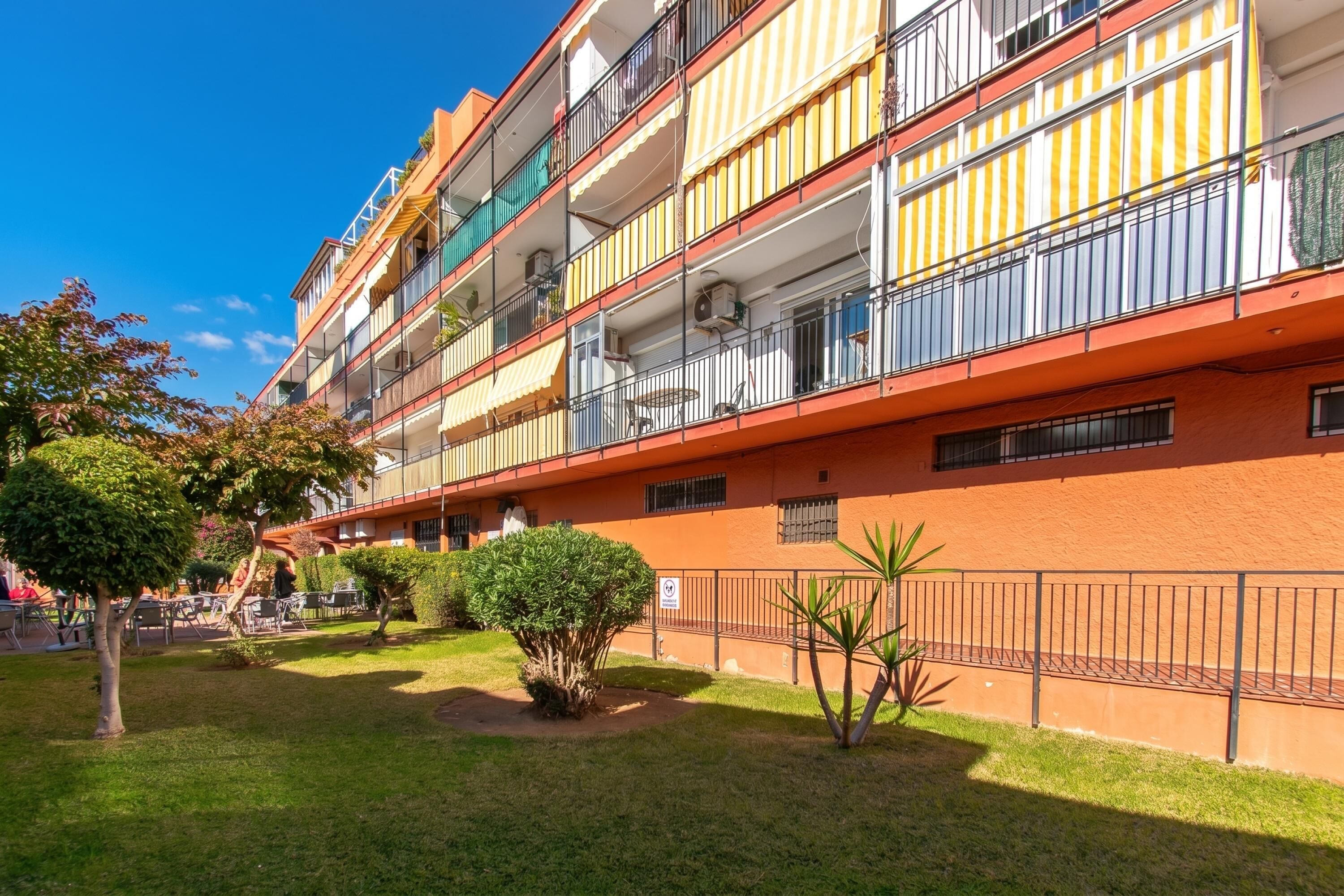 A multi-story building with balconies, a courtyard with grass and small trees, and a metal fence.