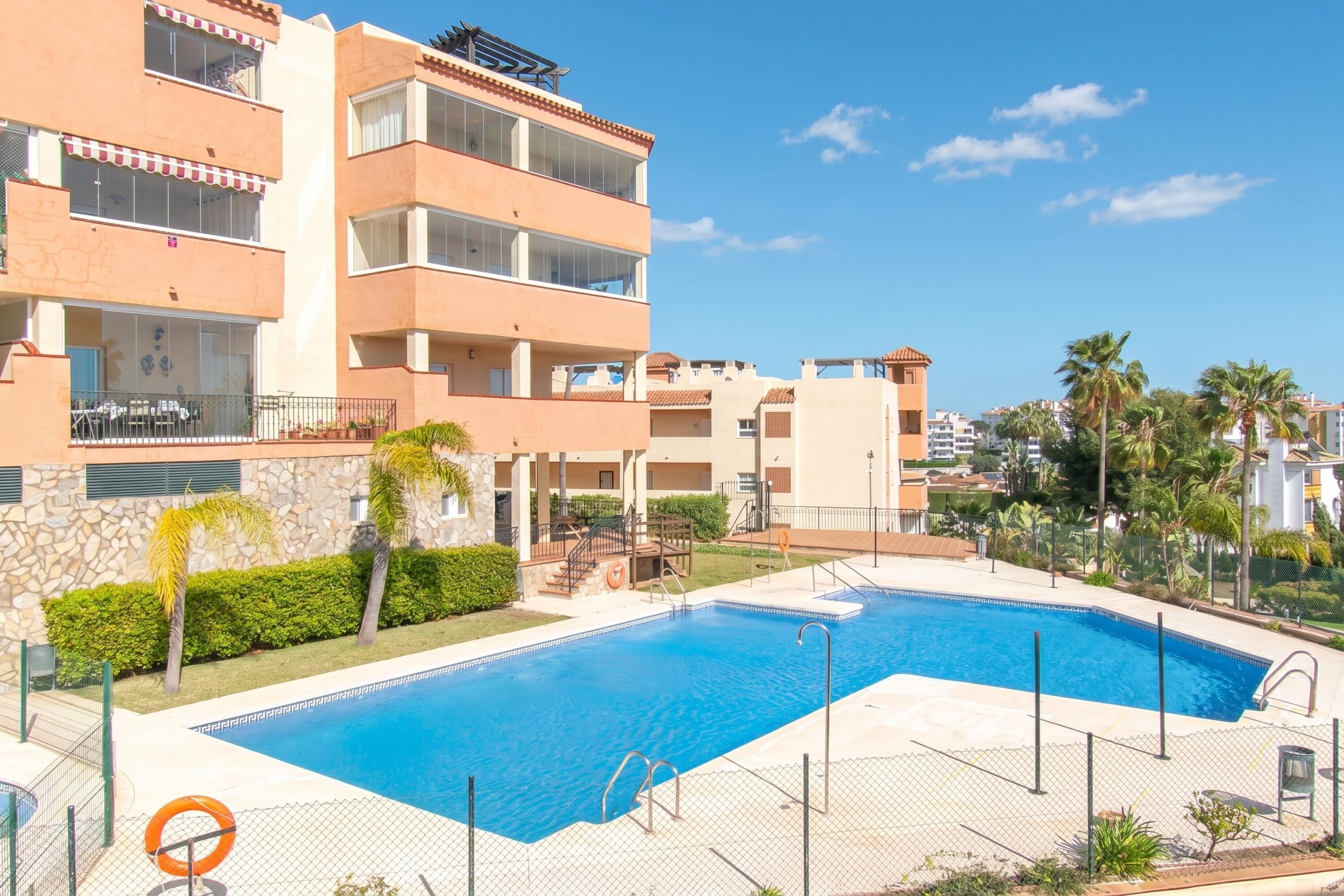 Une piscine entourée d’un immeuble à plusieurs étages doté de balcons, d’un mur en pierre et de palmiers.