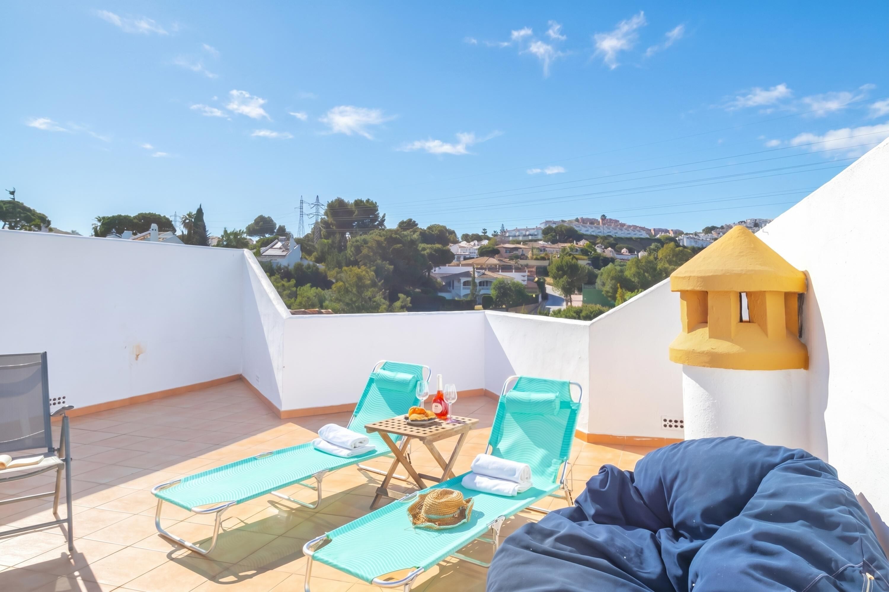 A rooftop terrace with two lounge chairs, a small table with a bottle and glasses, and a view of a town.