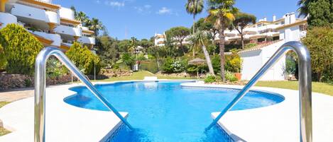 Una piscina con escaleras metálicas, rodeada de follaje y edificios blancos.