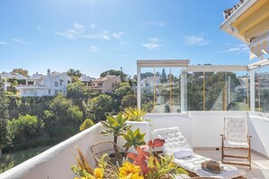A balcony with a glass enclosure, white cushioned seating, and a wicker chair. - Calahonda Golf (Mijas)
