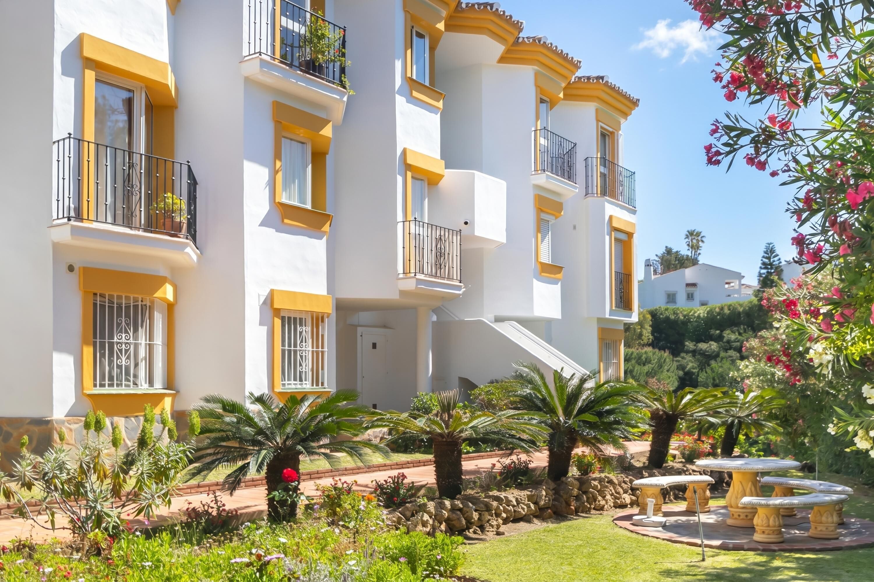 A multi-story building with balconies, a garden with palm trees, and a stone pathway.
